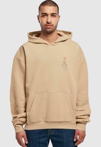 Felpa oversize beige in morbido misto cotone, dotata di tasca frontale e grafica di un cuore rosso sul petto a sinistra. Maniche lunghe con polsini a costine.