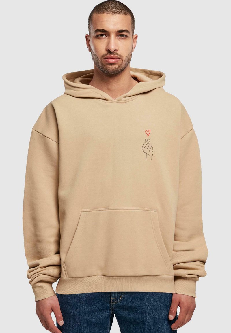 Felpa oversize beige in morbido misto cotone, dotata di tasca frontale e grafica di un cuore rosso sul petto a sinistra. Maniche lunghe con polsini a costine.