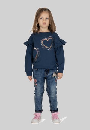 Marine sweatervest met ruches aan de mouwen, voorzien van gouden en kleurrijke hartontwerpen. Draag het met blauwe denim jeans, versierd met geborduurde patches, en roze schoenen.