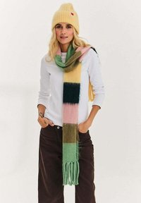 Kobieta w jasnopomarańczowej dzianinowej czapce, wielokolorowym paskowanym szaliku, białej koszuli z długim rękawem i ciemnobrązowych spodniach, z rękami w kieszeniach.