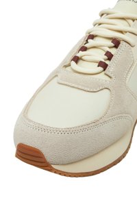Sneaker beige in suede con accenti in nylon crema, dotato di lacci tondi e occhielli marroni. Suola in gomma con scanalature orizzontali per una migliore aderenza.