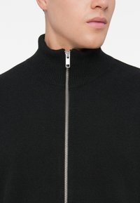 Mies, joka pitää yllä mustaa neulottua vetoketjullista neuletakkia, jossa on korkea kaulus. Vetoketju on osittain kiinni, paljastaen alaleuan ja kaulan valkoista taustaa vasten.