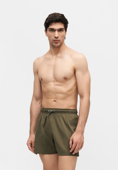 Jeune homme torse nu aux cheveux courts et foncés, portant un short vert olive à cordon, debout devant un fond gris clair uni.