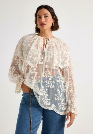 Lace blouse in cream with floral embroidery, layered design, and elastic cuffs; features tie closure at neck and light, translucent material.