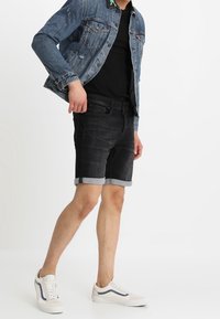 Giacca di jeans, pantaloni shorts in denim scuri con orlo arrotolato, top nero e sneakers bianche con accenti blu, tutti in posizione eretta.