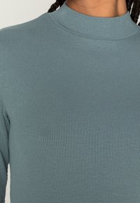 Nahaufnahme einer Person, die einen gerippten, langärmeligen Pullover mit hohem Kragen in gedämpftem Türkis trägt.