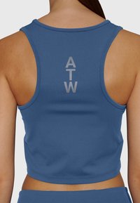 Kvinna som bär en blå racerback crop top med vertikala reflekterande bokstäver "ATW" centrerade på övre delen av ryggen mot en enfärgad bakgrund.