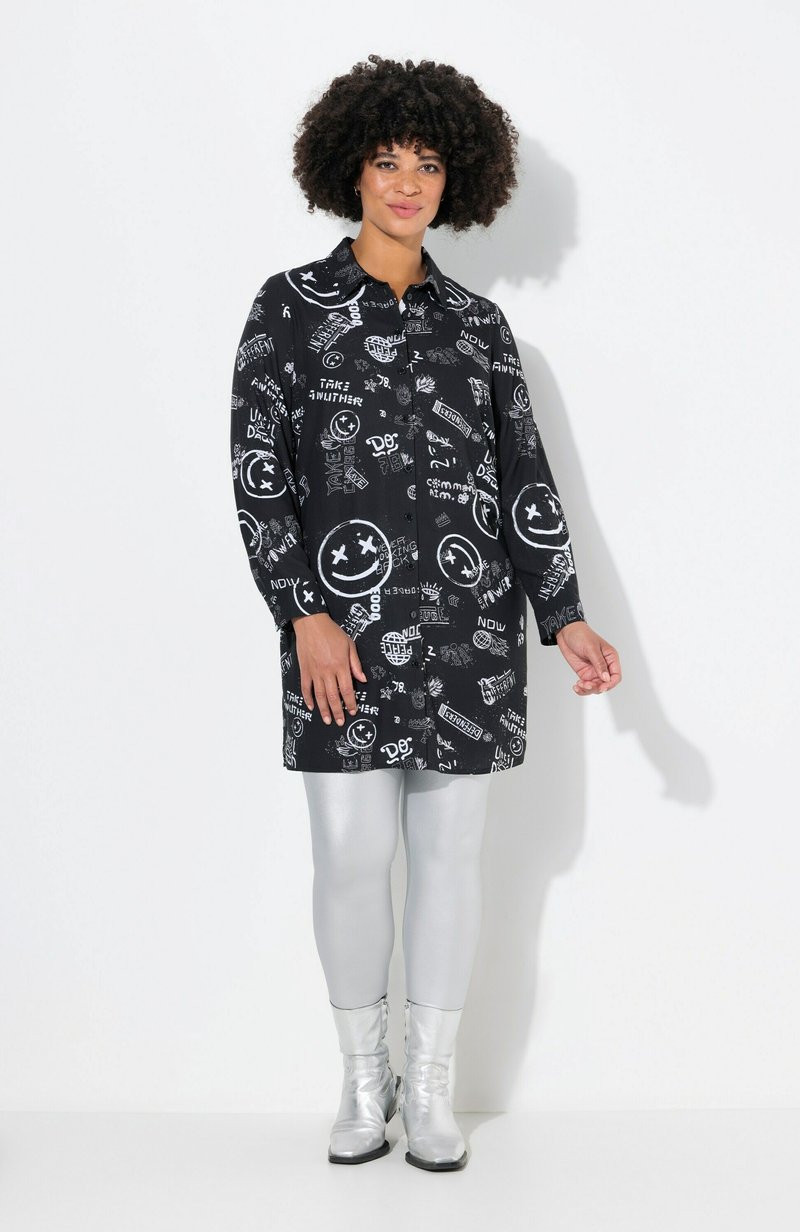 Vestido negro con patrones gráficos en blanco, con cuello tipo camisa, mangas largas y cierre delantero de botones, combinado con leggings plateados y botas.