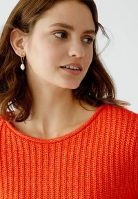 Helles orange gestricktes Sweatshirt mit weitem Ausschnitt und geripptem Muster, kombiniert mit goldenen Creolen, die eine weiße Perle enthalten.