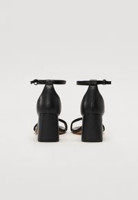 Paire de sandales noires en cuir à talon bloc avec brides à la cheville, vues de dos sur un fond clair uni.