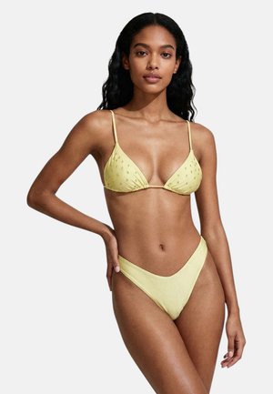 Donna con capelli lunghi e scuri che indossa un bikini giallo chiaro con puntini strutturati sulla parte superiore, posando con una mano sul fianco.