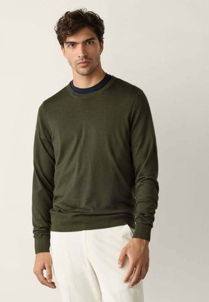 Homme aux cheveux courts et bouclés portant un pull vert foncé à manches longues par-dessus une chemise noire et un pantalon blanc, debout devant un fond uni.