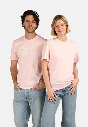 Homme et femme debout côte à côte portant des t-shirts "GERTRUDE" rose clair et des jeans bleu clair sur un fond clair uni.