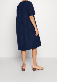 Navyblaues Baumwollkleid mit kurzen Ärmeln, rundem Ausschnitt und gerafftem Detail am Rücken. Getragen mit beigen geflochtenen Slippers.