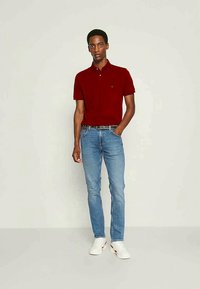 Červená polo košeľa s krátkymi rukávmi, modré deninové džínsy a biele tenisky. Polo košeľa má golier a malé logo; džínsy sú priliehavé.