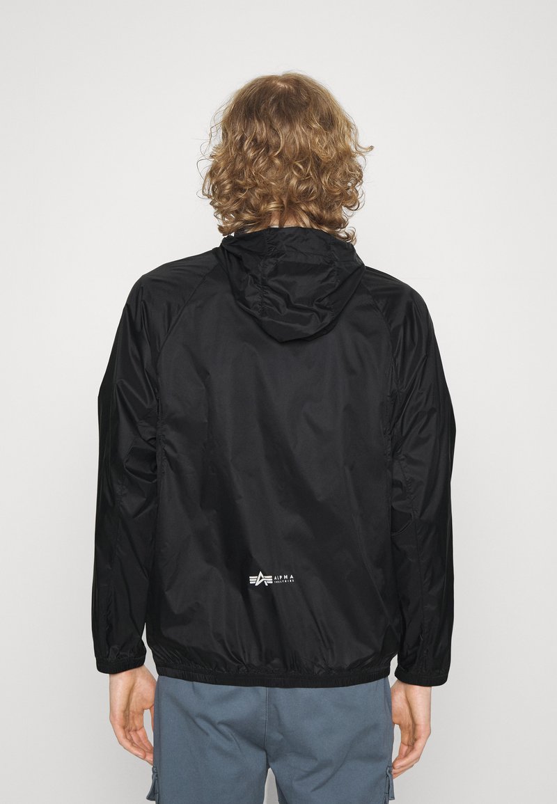 Schwarze, leichte Kapuzenjacke mit glatter Textur und elastischen Bündchen. Auf der Rückseite befindet sich ein einfaches Logo. Das Design ist minimalistisch.