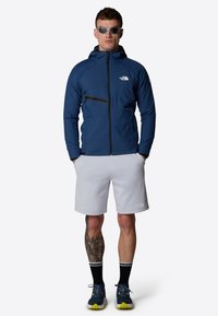 Marinblå lätt jacka med en dragkedjeficka, grå shorts och grå sneakers med gröna accenter. Solglasögon kompletterar outfiten.