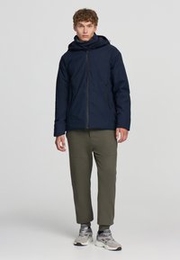Granatowa kurtka z kapturem na zamek, miękka w dotyku, z dwoma bocznymi kieszeniami; zestawiona z oliwkowymi dresami zwężanymi i szarymi trampkami.