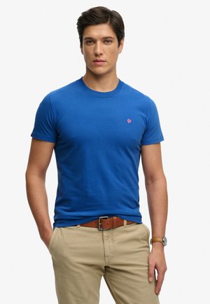 Hombre con camiseta azul con pequeño logo rojo, pantalones beige, cinturón marrón y reloj de pulsera, de pie con las manos parcialmente en los bolsillos.