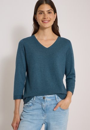 Femme souriante portant un pull à col en V manches longues couleur sarcelle et un jean bleu clair, la main droite dans la poche, sur un fond uni.