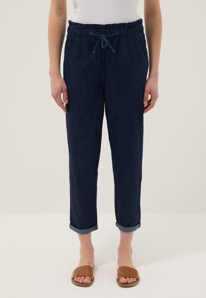 Vrouw die donkerblauwe, opgerolde broek met trekkoord draagt en bruine sandalen met open tenen, staand tegen een effen lichte achtergrond.