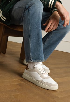 Witte sneaker met beige suède accenten en ton sur ton stiksel. Rond neus, low-top ontwerp, gecombineerd met opgerolde blauwe jeans en witte sokken.