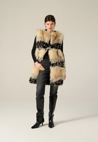 Gilet in pelliccia sintetica bianco e nero con un motivo geometrico, indossato sopra una maglietta nera, abbinato a jeans grigi e stivali al ginocchio neri lucidi.
