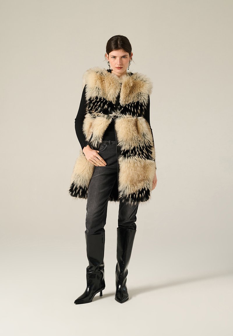 Gilet in pelliccia sintetica bianco e nero con un motivo geometrico, indossato sopra una maglietta nera, abbinato a jeans grigi e stivali al ginocchio neri lucidi.