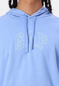 Šviesiai mėlyna džemperis su kepure, pagamintas iš minkštos medžiagos, su priekinėmis kišenėmis ir raišteliais, ant kurių yra "GAP" logotipas žaliais kontūriniais rašmenimis.