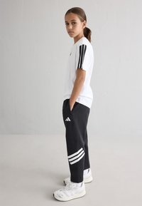 Baldais krekliņš ar melnām svītrām uz piedurknēm, melnas sporta bikses ar baltām akcentiem un baltas sporta kurpes. Gluda auduma tekstūra.