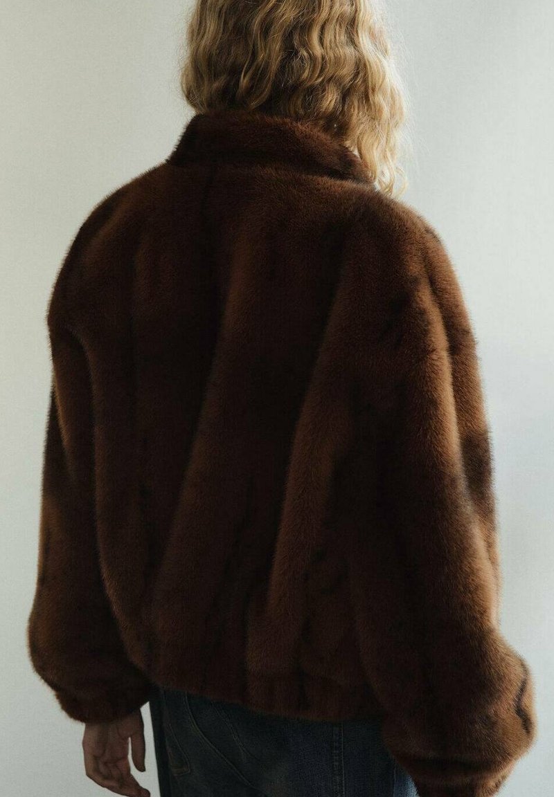 Personne aux cheveux blonds ondulés portant un manteau en fourrure peluche marron foncé, vue de dos sur un fond clair uni.