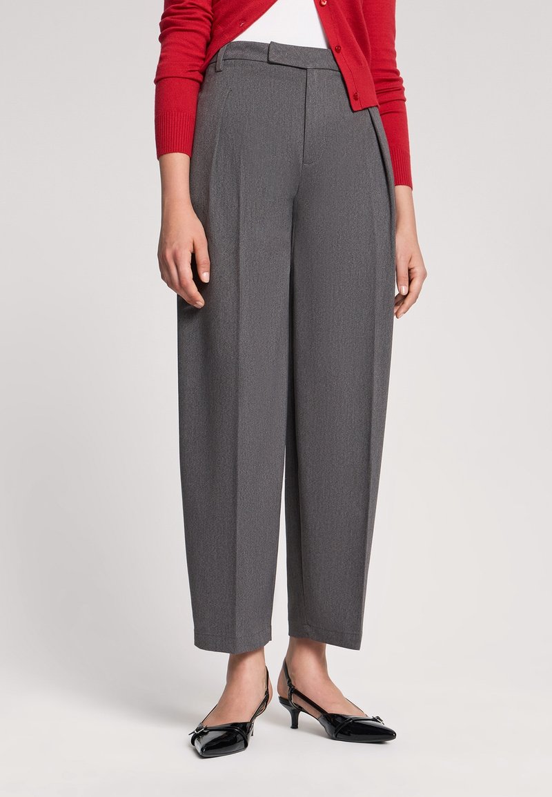 Donna con pantaloni grigi a vita alta e gamba larga, cardigan rosso, top bianco e scarpe slingback nere con tacco basso, in piedi su uno sfondo semplice.