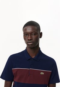 T-paita tummansinisellä pohjalla, jossa on viininpunainen vaakasuora raita ja valkoinen tehosteväri. Varustettu korkealla kauluksella ja vihreällä brodeeratulla logolla rinnassa.