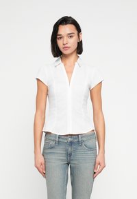 Λευκό, εφαρμοστό πουκάμισο με κοντά μανίκια, μυτερό γιακά και εμπρόσθιο φερμουάρ. Φορεμένο με ανοιχτόχρωμα τζιν σε τζην ύφασμα με κλασική γραμμή.