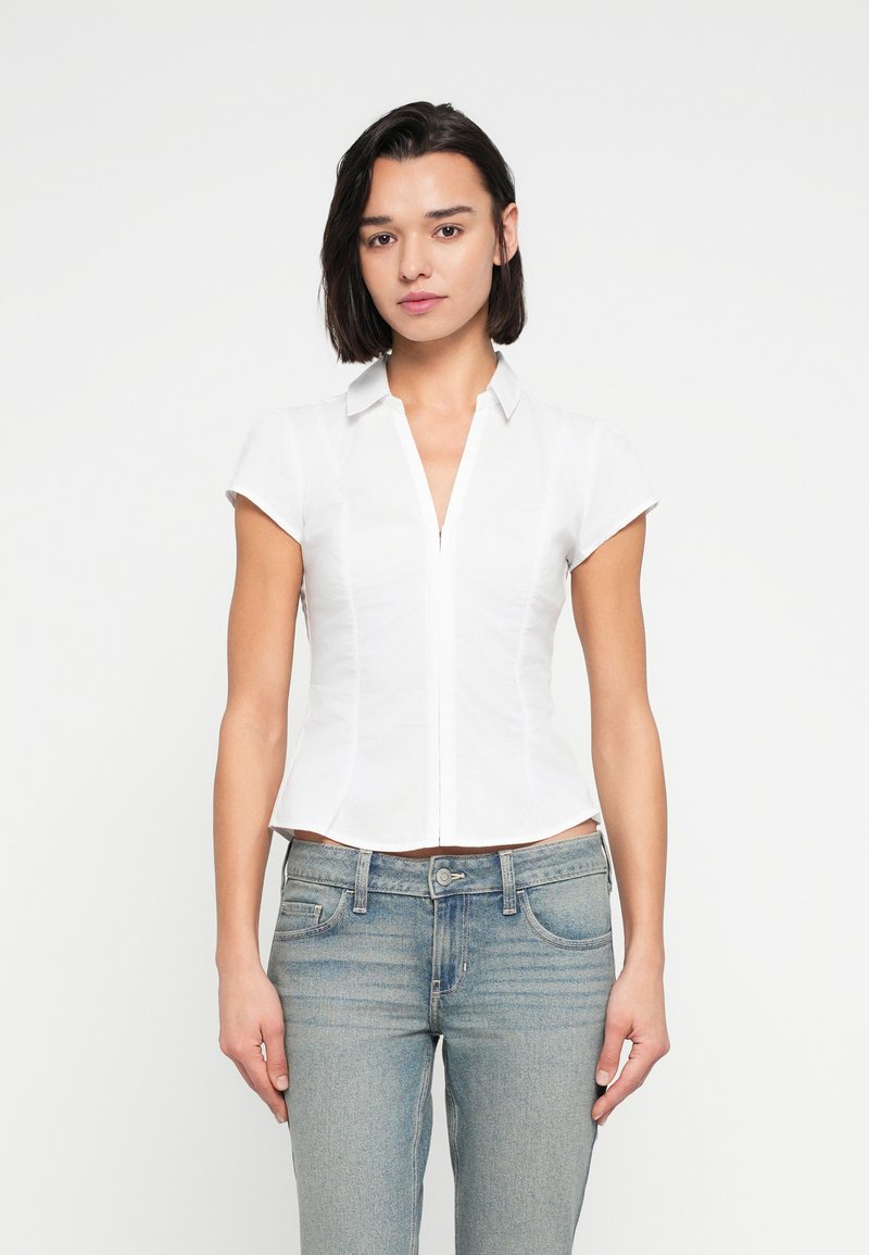 Λευκό, εφαρμοστό πουκάμισο με κοντά μανίκια, μυτερό γιακά και εμπρόσθιο φερμουάρ. Φορεμένο με ανοιχτόχρωμα τζιν σε τζην ύφασμα με κλασική γραμμή.