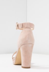 Béžová semišová sandál s vysokým podpatkem a kotníkovým páskem se zlatou sponou, zobrazená zespodu na bílé ploše a pozadí.