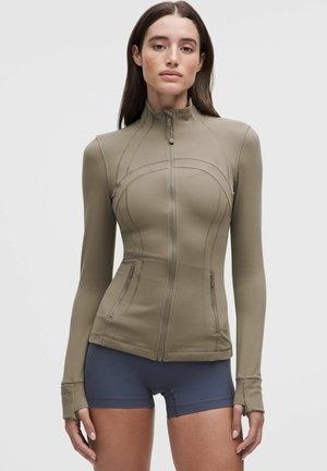 Vrouw draagt een aansluitend langarmig jasje met ritssluiting en duimgaten en donkere aansluitende korte broek tegen een effen lichte achtergrond.