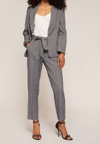 Šedý texturovaný oblek s dobře padnoucím sako a kalhoty s vysokým pasem na pásek. Model má na sobě černé sandály s pásky. Pod tím má bílý top.