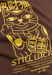 Κίτρινη γάτα maneki-neko με σηκωμένο πόδι και εικόνα νομίσματος σε καφέ ύφασμα, με κείμενο "ΠΑΡΑΜΕΝΩ ΤΥΧΕΡΟΣ" και περιγραφή από κάτω.