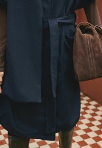 Person wearing a dark blue belted coat, brown long sleeves, brown boots, holding a large brown suede handbag on a red and white tiled floor.