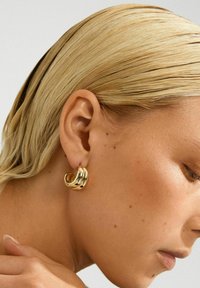 Nærbillede af en kvinde med slicked-back blond hår, iført en skinnende guld chunky hoop ørestik i sit venstre øre.