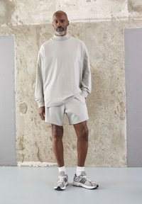 Svijetlosivi oversized dukserica s visokim ovratnikom, odgovarajuće šorts hlače i sivi sportski tenisici s bijelim detaljima. Teksturirana pozadina.