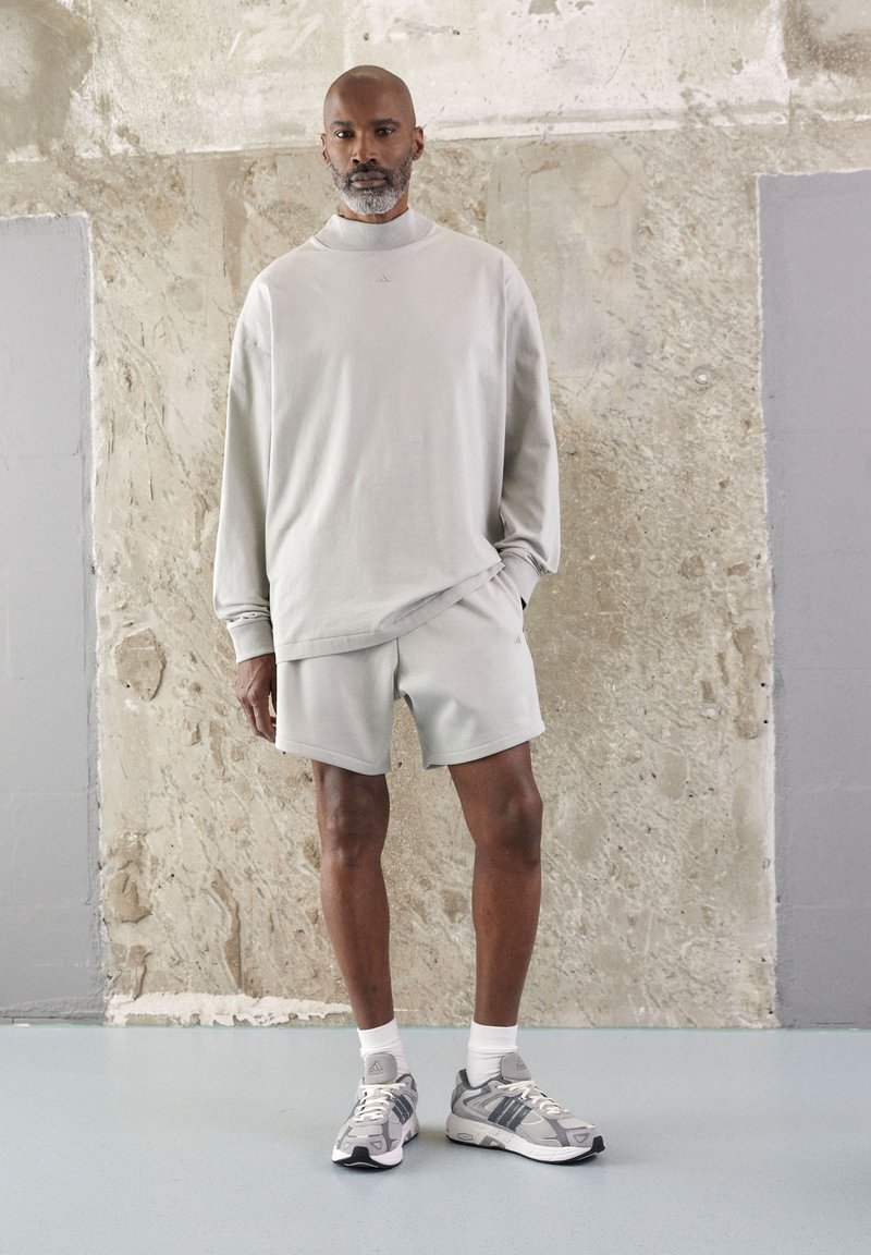 Sudadera gris claro de gran tamaño con un cuello alto, pantalones cortos a juego y zapatillas deportivas grises con acentos blancos. Fondo texturizado.