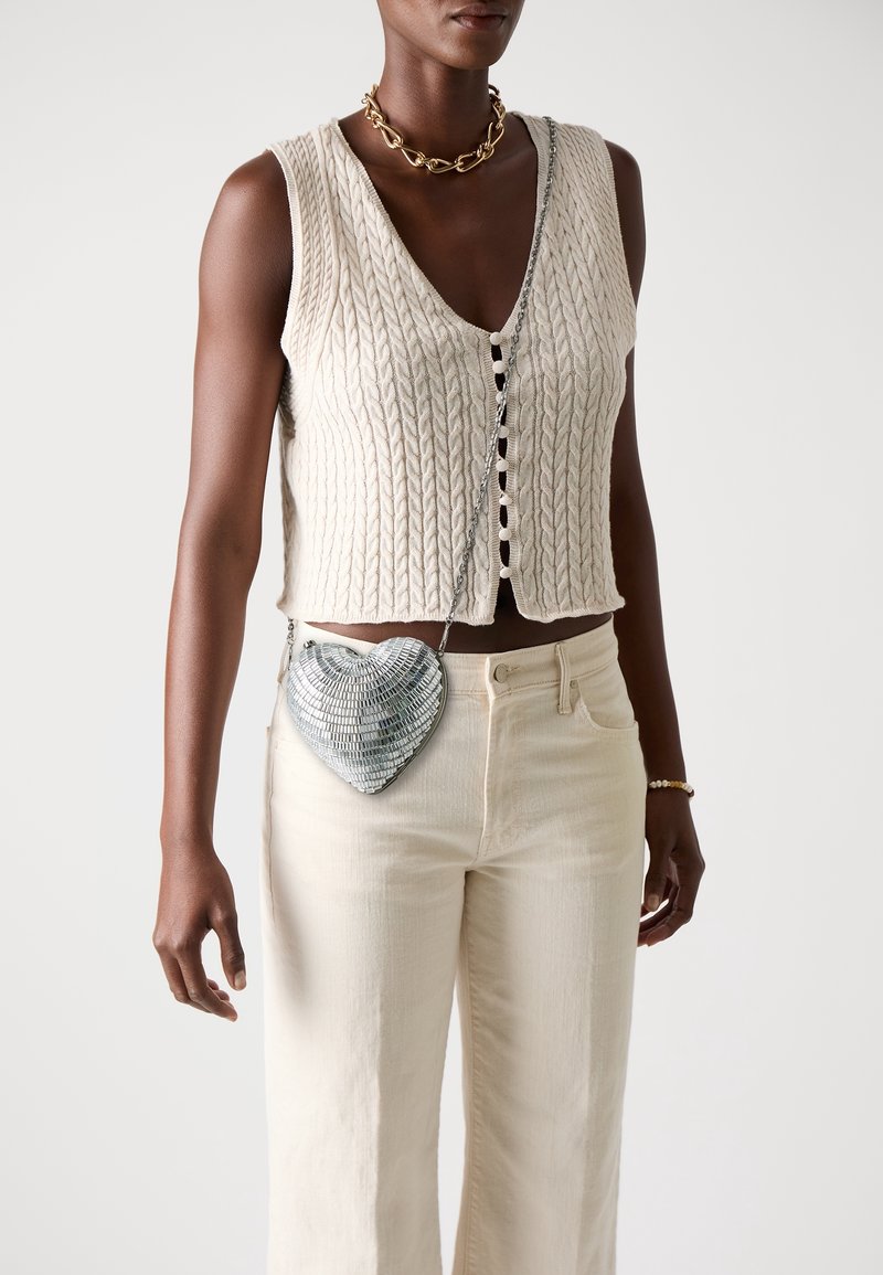 Personne portant un cardigan sans manches en maille crème, un pantalon crème, un collier en chaîne dorée et un petit sac en forme de cœur argenté avec une bandoulière chaîne.