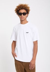 Jeune homme aux cheveux bouclés portant des lunettes de soleil noires, un t-shirt blanc Volcom et un pantalon en velours côtelé marron, debout avec une main dans la poche.