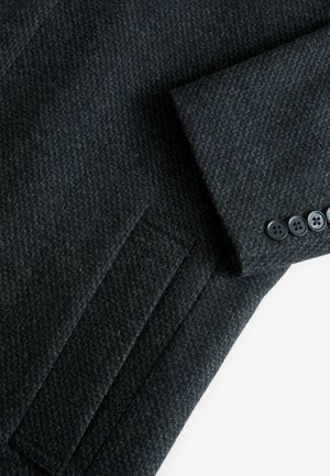 Donkergrijze wollen blazer met een gestructureerd herringbone patroon, gepofte schouders en zes zwarte knopenaccenten op de mouwen.