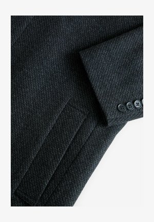 Sötétszürke gyapjú blézer, texturált halszálka mintázattal, párnázott vállrészekkel és hat fekete gombbal a mandzsettán.