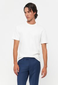 Mann mit schulterlangen dunklen Haaren, der ein schlichtes weißes kurzärmeliges T-Shirt und blaue Hosen trägt, vor einem weißen Hintergrund stehend.
