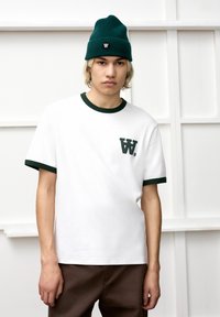 White cotton t-shirt with green ribbed collar and sleeve cuffs, featuring a small dark green logo on the left chest. Paired with brown pants.