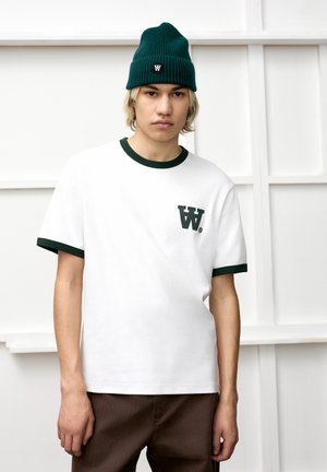 White cotton t-shirt with green ribbed collar and sleeve cuffs, featuring a small dark green logo on the left chest. Paired with brown pants.